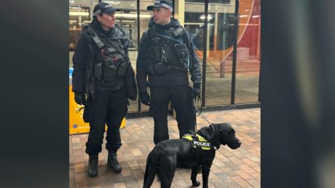 Police officers wearing black uniforms and black caps- standing in front of a black police dog with green jacket 