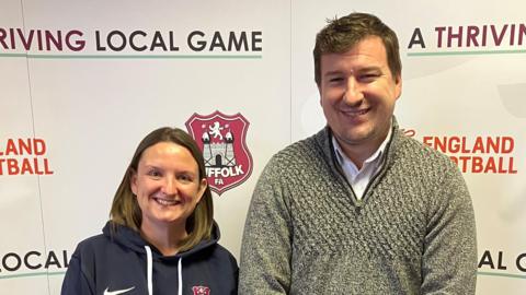 Hayley Chart and Andy Wilesmith from Suffolk FA standing side-by-side. They are both looking and smiling into the camera. There is advertising hoarding behind them.