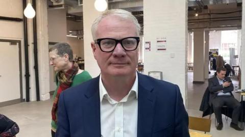 Richard Parker looks into the camera as he wears black-rimmed spectacles and a white shirt with a blue jacket over it. He has short, white hair, swept back. He stands in an office with people behind him and doorways. One man sits on a chair looking at a phone. Behind the mayor's right shoulder, a woman with short dark hair in a green jumper and multi-coloured scarf, looks to the side of the camera.