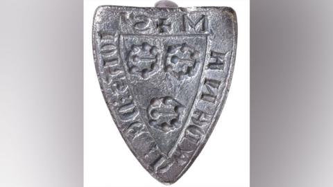 A silver medieval shield-shaped seal matrix from its front.  It has writing around its edge and in its middle are three brooch-shape designs, two above the third.