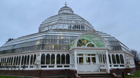 A close-up shot of the Palm House, a domed glass conservatory surrounded by parkland in Sefton Park.