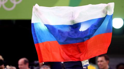 A man photographed from behind at the 2016 Olympics, arms outstretched holding a Russian flag behind his head