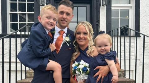 Leah is holding her 10 month old baby Hartlie. She is standing next to her partner Kyle who is holding their oldest child Jenson age- 4 in his arms. The family is all dressed in navy blue outfirs with Leah holding a bouquet of white and blue flowers.