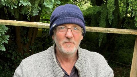 Man in a blue beanie wearing glasses with a beard and moustache with green foliage around 