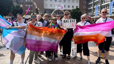 A group of eight women and one man pose for a picture outside in a city square, with high-rise civic buildings and others visible behind them. They hold up pink, orange and yellow rainbow flags and a sign saying Big Fat Lezza.
