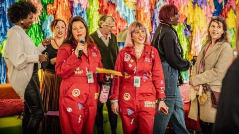 Two people waring red boiler suits covered in patches and images. The person on the left is speaking into a microphone. They are surrounded by a group of people and stood in front of a colourful, textile backdrop.