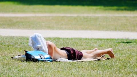 A man sunbathes on a patch of grass. He wears a pair of maroon shorts, with no top. Instead, what appears to be a white T-shirt is draped over his head, shielding his face from the sun. He lies on his front on a picnic blanket, with a blue rucksack next to him. 