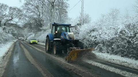 A tractor with snow plow on a snowy road with a police car behind