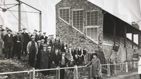 A black and white image of fans standing next to the stand at Borough Park in 1937, The stand is a brick construction with steps at the front leading up to the seats. The fans are standing on a mud bank. There are a few boys near the front while the rest are men all wearing hats.