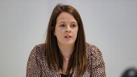 A woman with long brown hair sits in front of a thin black microphone. She wears a leopard print shirt, with a black top underneath. She has a thin silver necklace with a heart on it around her neck.