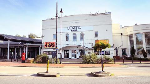 A large white building, with a sign saying Dorking Halls. There are trees and plants in front of the building. 