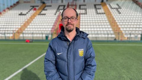 Jersey Football Association's chief executive Tim Pryor stands in front of an empty stand at a football pitch.