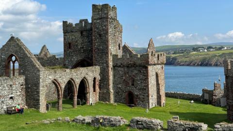A large part of the ruins of Peel Castle, which is built in sandstone and is surrounded by green grass with Peel Bay in the background with a headland above it.