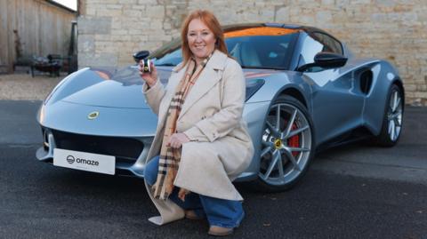 Claire Carty is crouched in front of a grey Lotus car. She has mid length red hair and is wearing a beige jacket and a checked scarf and brown boots and is holding up a set of car keys and smiling at the camera.