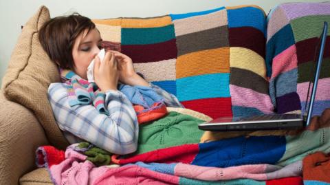 Woman wrapped in a colourful knitted blanket lying on a sofa with a laptop on her knees. She is blowing her nose into a hankerchief.