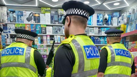A back view of three male police officers, wearing hi-vis yellow waistcoats with police written in white letters on blue backgrounds, over short-sleeved black tops and all wearing hats. They are facing a shop counter, beyond which are shelves packed a range of boxed goods.