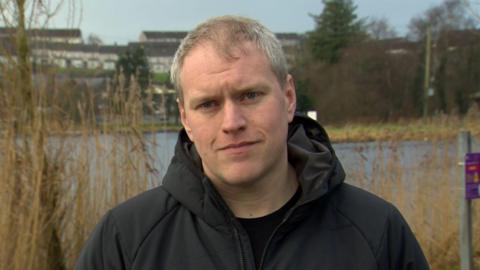 David is standing beside a lake. He's looking into the camera. It's an overcast day and he's wearing a black coat.