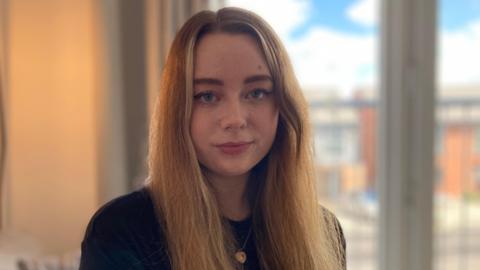 A young white woman with long medium blonde hair and blue eyes is looking straight at the camera. She is wearing a black top with a gold necklace around her neck. There is a lamp to the left of the picture and windows to the right.