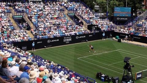 A crowd of people sitting in stands watching tennis - the green grass tennis court is in the centre of the image