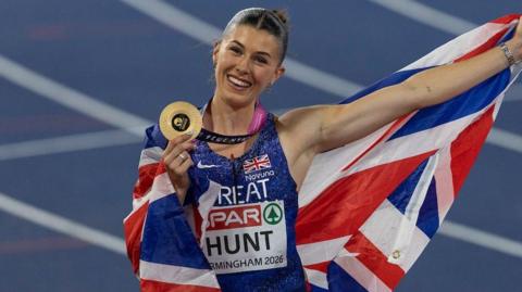 Amy Hunt - a woman in a sparkly blue Nike vest - is draped in a union jack flag which she is holding out into the air with one hand. In her other hand she is holding a gold circular medal. She has brown hair slicked back and is smiling. A blurred racetrack can be seen behind her.