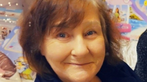 The picture shows a woman sitting indoors, surrounded by bright and colourful items. They are wearing a dark top.