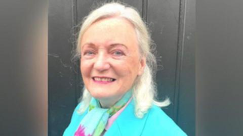 A woman with blond shoulder length hair and pink lipstick smiling . She is turned towards the camera but her shoulders are angled away. The shoulders of her turquoise jacket can e seen and she is wearing a turquoise, pink and green neck scarf. A dark grey fence can be seen behind her.