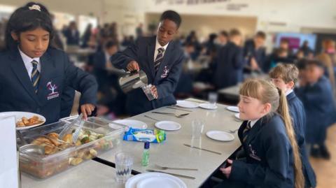 Four students wearing school uniforms sit and stand around a table. A girl is serving potatoes and a boy is pouring water. There is a room full of students in the background. 