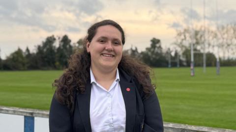 A woman with long brown curly hair, wearing a white blouse and dark blue jacket standing beside a grassy rugby pitch
