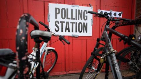 Two bikes are chained up outside a polling station in front of a bright red wall. 