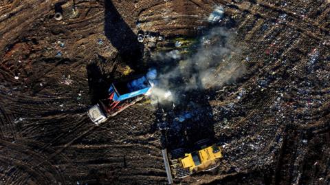 An aerial image showing two vehicles, one blue, one yellow, working on a landfil with smoke rising from the site.