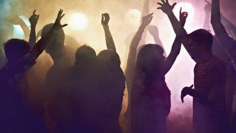 Silhouette of people dancing in a club with green, yellow and pink lights and smoke behind.