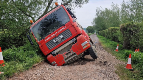 A red lorry leaning to the right. One wheel is in a hole on an uneven surface. There is a long road behind it with trees and hedges either side. The writing on the lorry says "stabilised pavements".