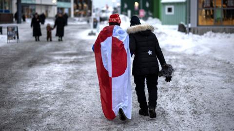 Two people walk down an icy street. One is wearing a draped Greenland flag.