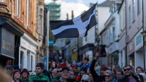 A large black-and-white Cornish flag is held above a crowd on a busy town street lined with historic buildings for Redruth's St Piran's Festival.
