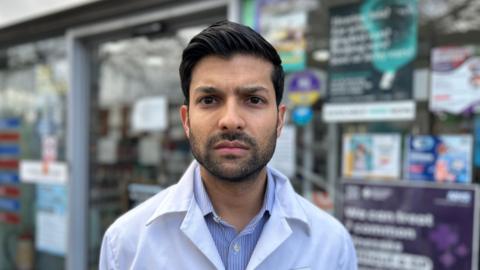 A young man with short hair and a beard is wearing a white coat. He is looking stern and standing in front of a shop front. 