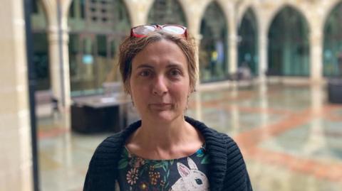 Tina Matthew with short brown hair wearing glasses on her forehead and a blue cardigan with blue dress.  There is a picture of a rabbit on the dress. She is standing in the pillared stone courtyard of Northampton Guildhall.