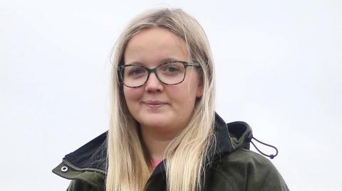 Eveey Hunter has long blonde hair and is wearing a dark green waterproof coat as she looks at the camera on an overcast day.