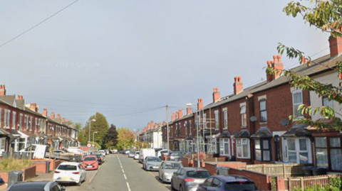 Residential street lined with red-brick terraced houses on both sides, with cars parked along the road. A clear roadway extends into the distance beneath an overcast sky, with trees, utility lines, and scaffolding visible among the houses.
