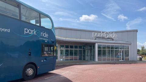 On the left hand side there is a blue double decker bus, on the left there is a silver coloured building with the words 'Park and Ride' at the front. Portsmouth Park and Ride.