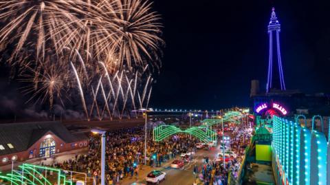 Aerial view of the fireworks display on the beach near Blackpool's North pier showing crowds of people watching on the promenade in the evening with the Blackpool Illuminations on and Blackpool Tower lit up purple in the background.