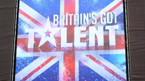 A large billboard poster showing the Britain's Got Talent logo, which is white writing with a star for the letter 'A' in the word talent, against a backdrop of a sparkly union flag