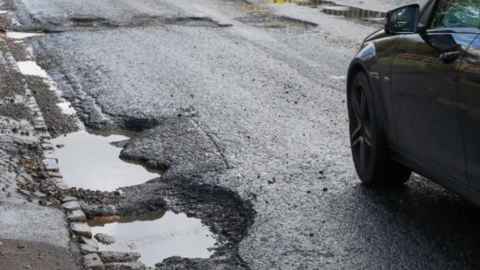 Two potholes at the side of the road are in the bottom left of the photo and others are in the middle of the road a few yards ahead. Part of a car is on the far right of the image.
