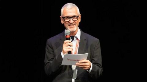 William Burdett-Coutts, a man with grey hair, is wearing glasses and a black suit with white shirt and orange lanyard. He is holding a microphone and a piece of paper, and smiling as he looks off to the right hand side of the frame.