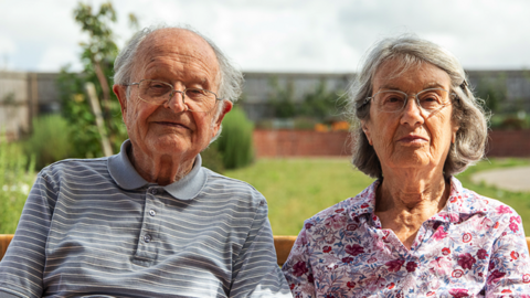 Peter and Shirley sit on a garden bench with lots of green shrubs in the background. Peter is wearing a grey striped polo shirt and Shirley is wearing a red flower patterned top.
They are holding hands. 
Both have grey hair and are aged in their 90s. 