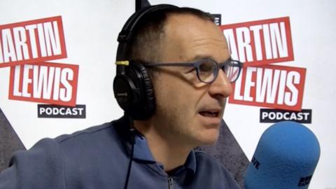 Martin Lewis wearing headphones and speaking in front of a microphone. There is branding for the Martin Lewis Podcast in red and white in the background.