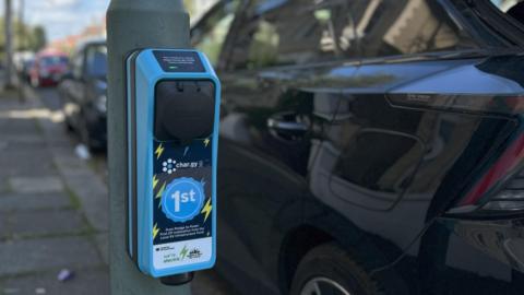An electric charger attached to a street lamppost on a pavement next to a parked car