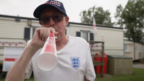 Richard blows an England themed vuvuzela trumpet, wearing a blue England Cap, white England shirt and sunglasses. Behind him is his static caravan decorated with England flag bunting.
