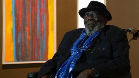 Sir Frank Bowling looks at the camera unsmiling. He is in a wheelchair and behind him is a brightly coloured orange and red abstract painting. Sir Frank is wearing a black hat and jacket and blue scarf.
