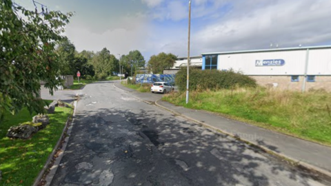 A white industrial unit at Westmorland Business Park in Kendal. There is a worn single track road with potholes skirting around the site. There is green grass and trees on either side of the road.