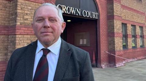 Derek Bennett: a grey haired man wearing a grey jacket and red tie outside a red-and-brown brick building. The words "crown court" are written above the entrance to the building in capital letters.